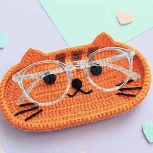 May include: An orange crocheted cat-shaped tray with dark brown stripes, black embroidered eyes, a nose, and whiskers. A pair of clear-framed spectacles rests on top of the tray, which features two small pointed ears at the top edge.