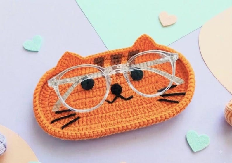 May include: An orange crocheted cat-shaped tray with dark brown stripes, black embroidered eyes, a nose, and whiskers. A pair of clear-framed spectacles rests on top of the tray, which features two small pointed ears at the top edge.
