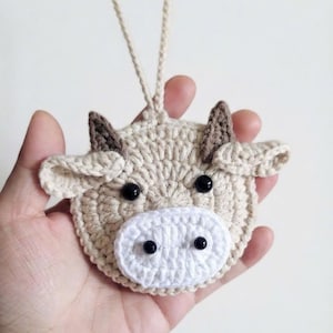 Op de afbeelding: Een rond, beige gehaakt ornament in de vorm van een koeienkop met twee bruine puntige oren, twee brede beige oren, een witte ovale snuit en vier kleine zwarte kraaloogjes. Een beige gehaakt koordje is aan de bovenkant bevestigd om op te hangen.