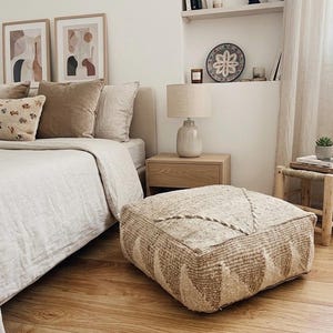 Op de afbeelding: Een slaapkamer met een bed met neutrale lakens en kussens. Een geweven poef met een geometrisch patroon staat op de houten vloer. Een nachtkastje met een lamp en kunstwerken maken de ruimte gezellig.