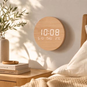 Può includere: Un orologio digitale rotondo in legno chiaro che mostra l'ora 10:08, la data e la temperatura. L'orologio è montato su una parete bianca. Il quadrante dell'orologio ha un motivo a venatura di legno chiaro. L'ora, la data e la temperatura sono visualizzate in bianco.