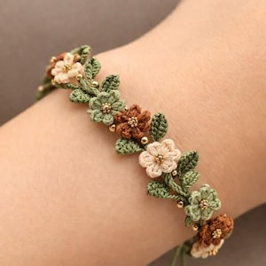 Könnte beinhalten: Gehäkeltes Armband mit floralem Design. Das Armband hat grüne Blätter und Blüten in Braun-, Creme- und Grüntönen. Kleine goldene Perlen akzentuieren die Blüten. Das Armband ist mit einer grünen Kordel gebunden.