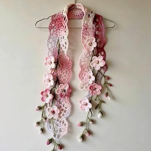 Könnte beinhalten: Handgefertigter Häkelschal in Rosa, Weiß und Lavendel, verziert mit dreidimensionalen Blumen. Der Schal wird auf einem silbernen Kleiderbügel präsentiert, der sein filigranes Spitzenmuster und die floralen Akzente hervorhebt.