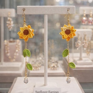 May include: A pair of handmade sunflower earrings. Each earring features a yellow and brown crocheted sunflower, green crocheted leaves, and a clear crystal bead. The earrings hang from gold-colored hooks and chains. The sign below reads "Sunflower Earrings."