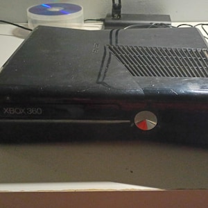 May include: Black Xbox 360 console with a silver power button and a disc tray. The console has a vent on top and the text "XBOX 360" is visible on the front. A CD is visible in the background.