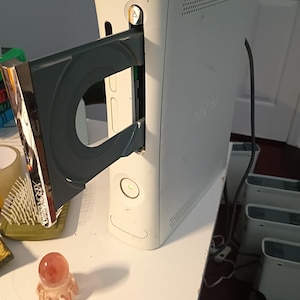 May include: A white Xbox 360 video game console standing vertically with its disc tray extended. The console features a power button with a green light, a chrome-coloured disc tray faceplate with the Xbox 360 logo, and a black power cable.