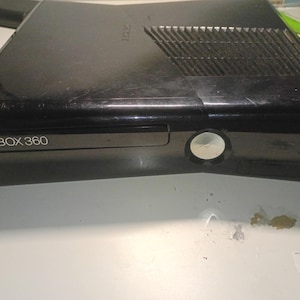 Könnte beinhalten: Schwarze Xbox 360 Konsole mit dem Schriftzug "XBOX 360" auf der Vorderseite. Sie verfügt über ein Laufwerk, einen Power-Knopf und Lüftungsschlitze. Die Oberfläche weist Gebrauchsspuren auf.