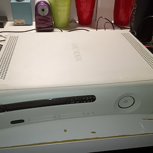 Könnte beinhalten: Eine weiße Xbox 360 Videospielkonsole mit einem rechteckigen Gehäuse, einem Laufwerksschlitz, einem runden Einschaltknopf und Lüftungsgittern auf der Oberseite. Das Xbox 360 Logo ist auf der oberen Gehäuseplatte eingeprägt.