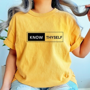 Puede incluir: Una camiseta de manga corta color amarillo mostaza con un gráfico rectangular negro en el pecho que contiene el texto blanco "KNOW" y el texto negro "THYSELF" dentro de un recuadro blanco. La prenda tiene cuello redondo y mangas dobladas.