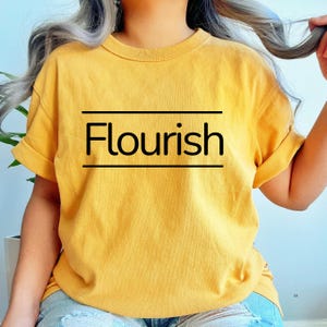May include: A mustard yellow short-sleeve t-shirt featuring the word Flourish printed in black, centered between two horizontal black lines. The shirt has a crew neckline and rolled sleeves, providing a casual look with a clear graphic design.