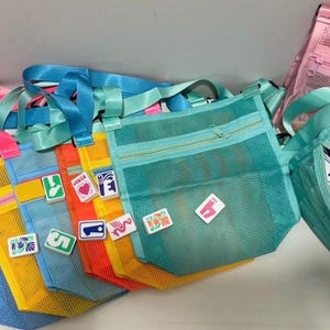 May include: A collection of colorful mesh tote bags with matching straps. The bags are in shades of pink, blue, yellow, orange, and teal. Some bags have decorative patches. One bag is open, revealing game tiles.