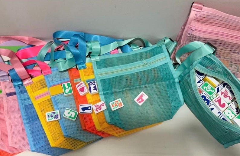 May include: A collection of colorful mesh tote bags with matching straps. The bags are in shades of pink, blue, yellow, orange, and teal. Some bags have decorative patches. One bag is open, revealing game tiles.