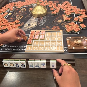 May include: A Mahjong game in progress, featuring a dark game board with a floral and insect design. The game includes numerous tiles, a wooden dice tray with two dice, and a tile rack. The image shows hands arranging the tiles.