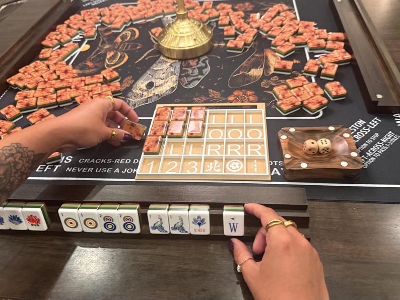May include: A Mahjong game in progress, featuring a dark game board with a floral and insect design. The game includes numerous tiles, a wooden dice tray with two dice, and a tile rack. The image shows hands arranging the tiles.