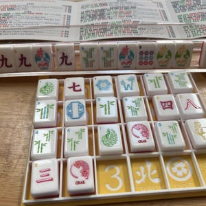 May include: A Mahjong game set with white tiles featuring various symbols and characters in red, green, and blue. The tiles are arranged in a yellow tray, with a printed guide in the background.