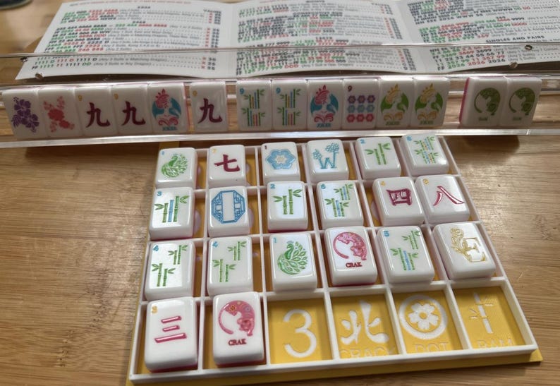 May include: A Mahjong game set with white tiles featuring various symbols and characters in red, green, and blue. The tiles are arranged in a yellow tray, with a printed guide in the background.