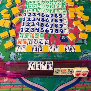 May include: A complete Mahjong set with tiles featuring numbers and symbols in various colors. The set includes tiles with the word "Joker" and the word "News". The game is set on a floral patterned mat.