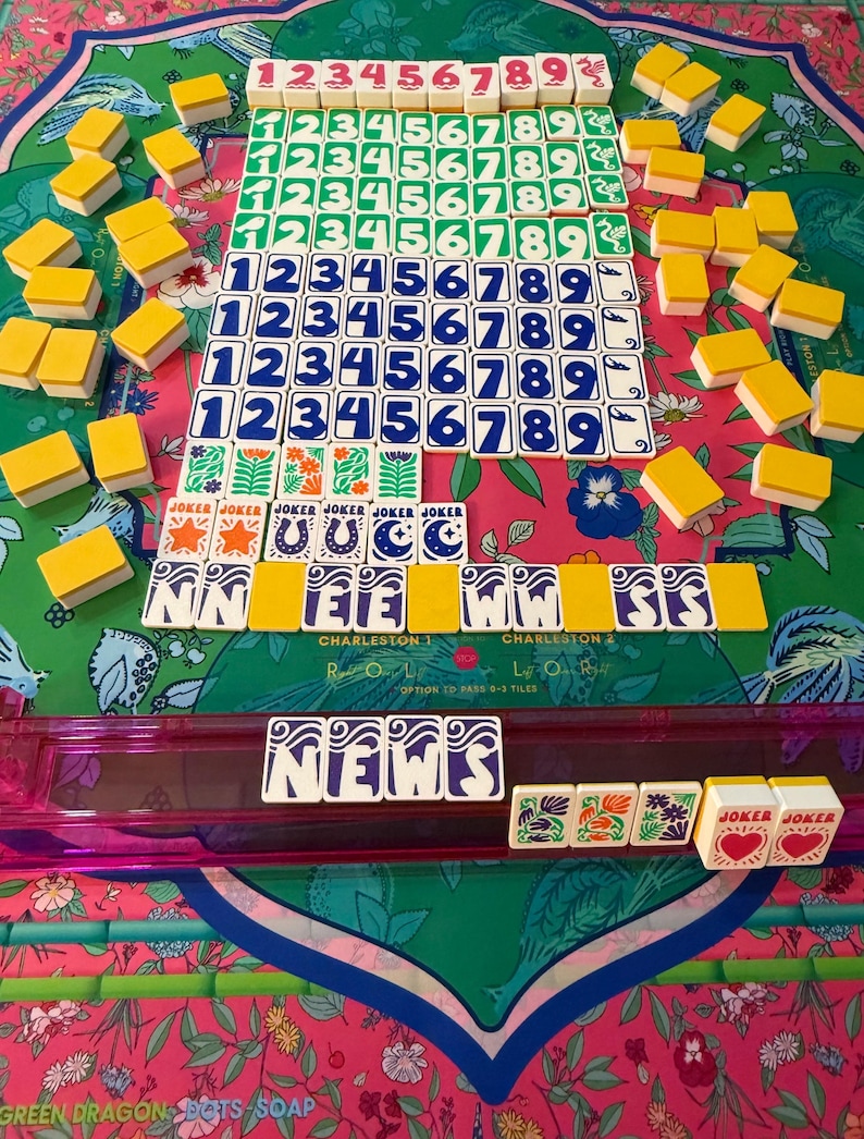 May include: A complete Mahjong set with tiles featuring numbers and symbols in various colors. The set includes tiles with the word "Joker" and the word "News". The game is set on a floral patterned mat.
