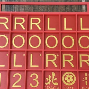 May include: A red game board with a grid of squares, each containing a letter, number, or symbol in gold. The letters include R, L, and O. Numbers 1, 2, and 3 are also present, along with symbols. The board has a handle at the top.
