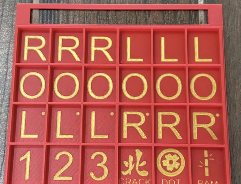 May include: A red game board with a grid of squares, each containing a letter, number, or symbol in gold. The letters include R, L, and O. Numbers 1, 2, and 3 are also present, along with symbols. The board has a handle at the top.