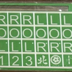 May include: A green and white game board with a grid of letter and number tiles. The tiles display the letters R, O, and L, along with the numbers 1, 2, and 3. The game board also includes symbols and characters.