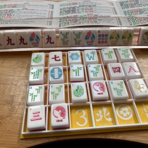 May include: A Mahjong set with white tiles featuring various symbols and characters in red, green, and blue. The tiles are arranged in a yellow tray, with a wooden surface in the background. A rule sheet is visible.