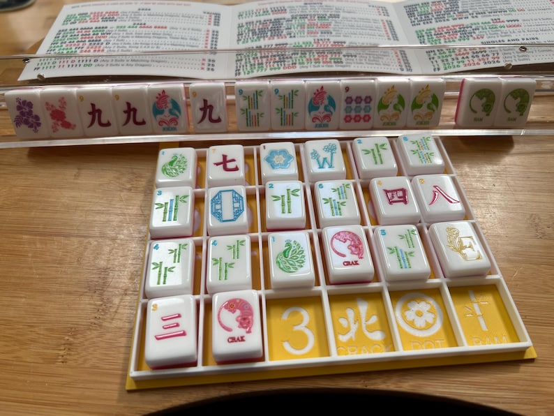 May include: A Mahjong set with white tiles featuring various symbols and characters in red, green, and blue. The tiles are arranged in a yellow tray, with a wooden surface in the background. A rule sheet is visible.