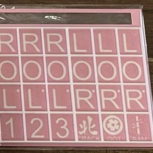 May include: A pink Scrabble tile set with white letter tiles. The tiles spell out the words "ROLL" and "RACK DOT RAM" with numbers and symbols. The set is in a clear plastic package.