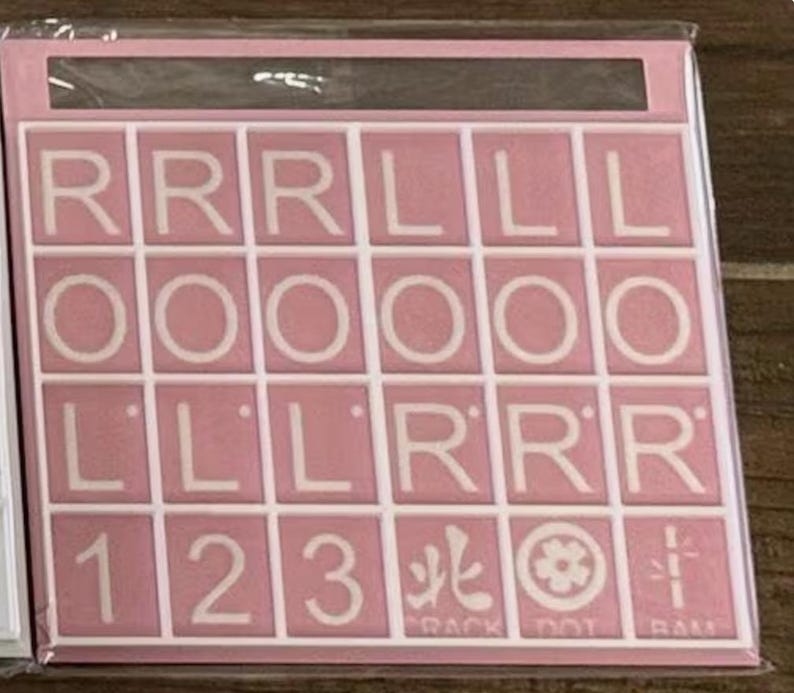 May include: A pink Scrabble tile set with white letter tiles. The tiles spell out the words "ROLL" and "RACK DOT RAM" with numbers and symbols. The set is in a clear plastic package.