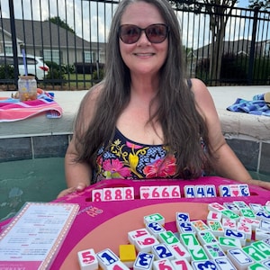 May include: A woman in a floral swimsuit smiles while playing a board game. The game features a pink board with tiles displaying numbers and symbols. The game is called "Debuta".