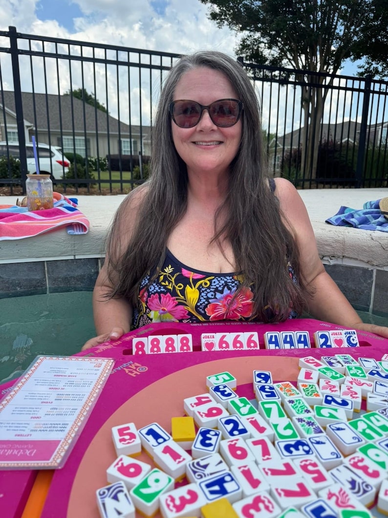 May include: A woman in a floral swimsuit smiles while playing a board game. The game features a pink board with tiles displaying numbers and symbols. The game is called "Debuta".