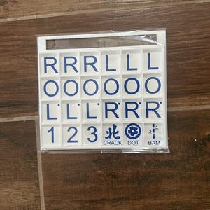 May include: A white plastic tile set with blue lettering and symbols. The tiles are arranged in a grid and include letters, numbers, and Chinese characters. The words "CRACK DOT BAM" are printed at the bottom.