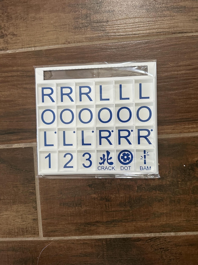 May include: A white plastic tile set with blue lettering and symbols. The tiles are arranged in a grid and include letters, numbers, and Chinese characters. The words "CRACK DOT BAM" are printed at the bottom.
