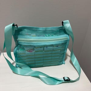 May include: A turquoise mesh crossbody bag with a zippered top and adjustable strap. The bag is filled with various items, and the strap is secured with a black buckle. The bag is sitting on a white surface.