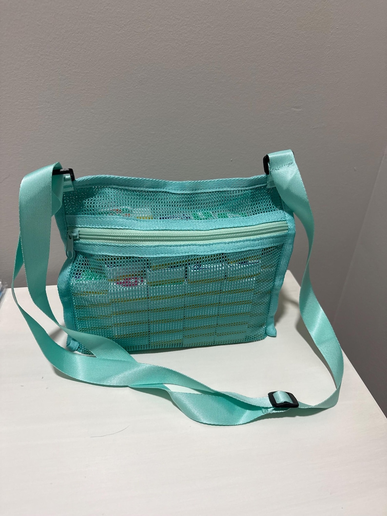 May include: A turquoise mesh crossbody bag with a zippered top and adjustable strap. The bag is filled with various items, and the strap is secured with a black buckle. The bag is sitting on a white surface.