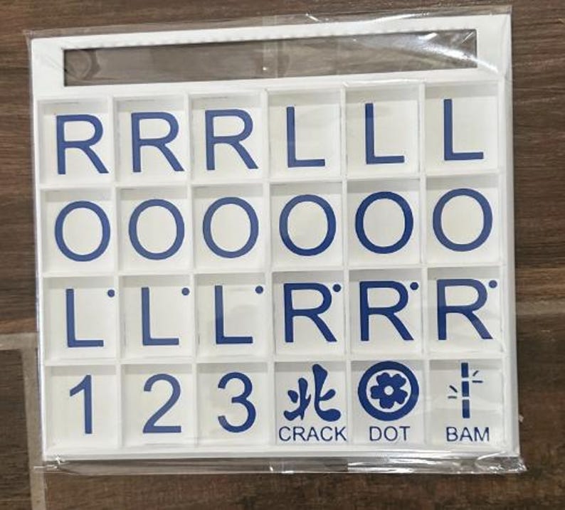 May include: A white game tile set with blue lettering and symbols. The set includes letter tiles with the letters R, L, and O, along with number tiles 1, 2, and 3. The set also includes tiles with the words CRACK, DOT, and BAM.