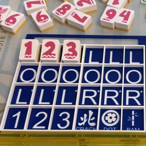 May include: A close-up of a Mahjong set with white tiles featuring pink numbers and symbols. The set includes a blue and white tile holder with the numbers 1, 2, and 3. The word "JOKER" is visible on one tile.