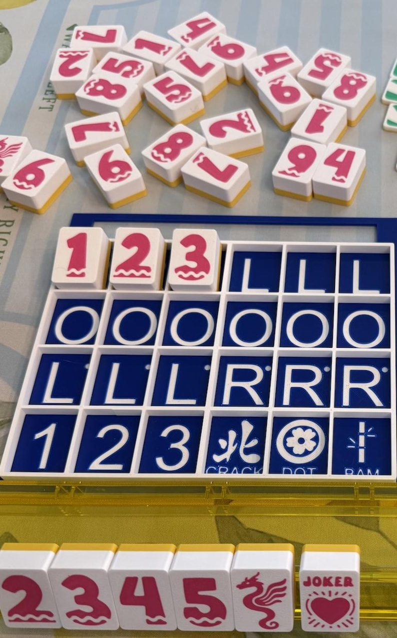 May include: A close-up of a Mahjong set with white tiles featuring pink numbers and symbols. The set includes a blue and white tile holder with the numbers 1, 2, and 3. The word "JOKER" is visible on one tile.