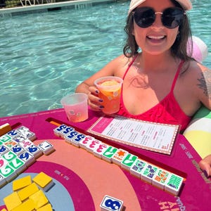 May include: A woman wearing a white cap with "MAHJ SQUAD" in blue text, and sunglasses, is in a pool. A pink and orange Mahjong game board with tiles is in front of her. A drink with a slice of orange is in her hand.