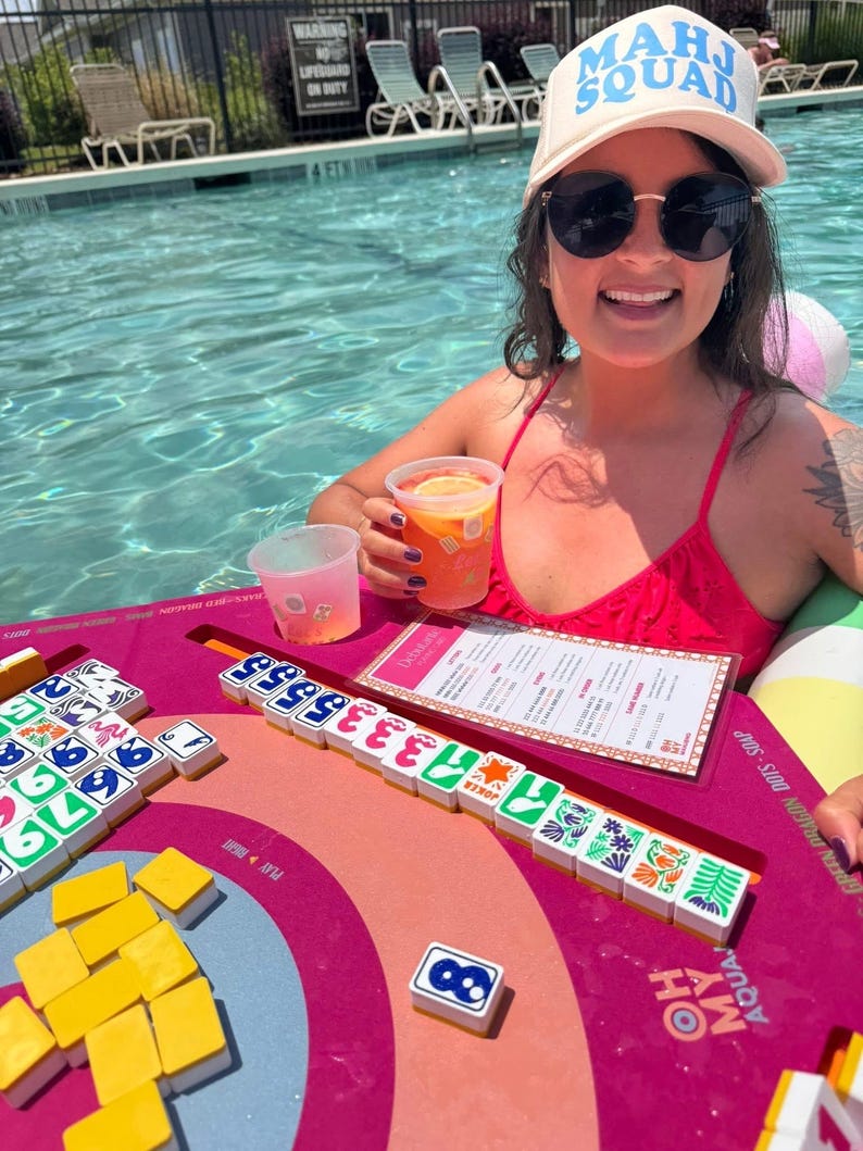 May include: A woman wearing a white cap with "MAHJ SQUAD" in blue text, and sunglasses, is in a pool. A pink and orange Mahjong game board with tiles is in front of her. A drink with a slice of orange is in her hand.