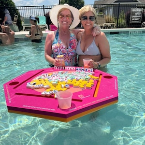 May include: Two women in a swimming pool with a floating pink game board. The board is filled with game tiles. The women are wearing sunglasses and swimsuits. The water is clear and blue.