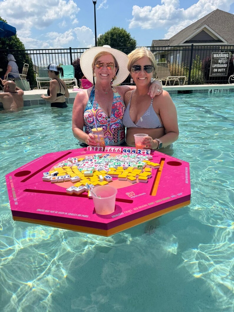 May include: Two women in a swimming pool with a floating pink game board. The board is filled with game tiles. The women are wearing sunglasses and swimsuits. The water is clear and blue.