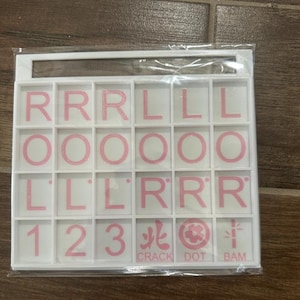 May include: A white game tile set with pink letters, numbers, and symbols. The tiles include the letters R, L, and O, the numbers 1, 2, and 3, and the words CRACK, DOT, and BAM. The tiles are arranged in a grid.