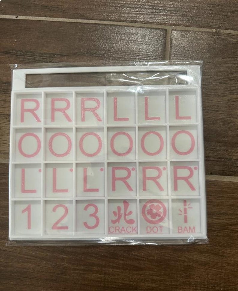 May include: A white game tile set with pink letters, numbers, and symbols. The tiles include the letters R, L, and O, the numbers 1, 2, and 3, and the words CRACK, DOT, and BAM. The tiles are arranged in a grid.
