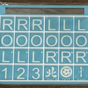 May include: A Scrabble tile set with white letter tiles on a light blue background. The tiles spell out the word "ROLL" multiple times, along with numbers and symbols. The set is contained within a light blue frame.