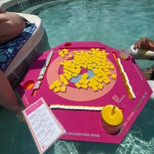 May include: A pink hexagonal floating game board with yellow tiles and game pieces. The board is in a swimming pool with people in the water. A menu is visible, along with the text "OH MY AQUAJONG".
