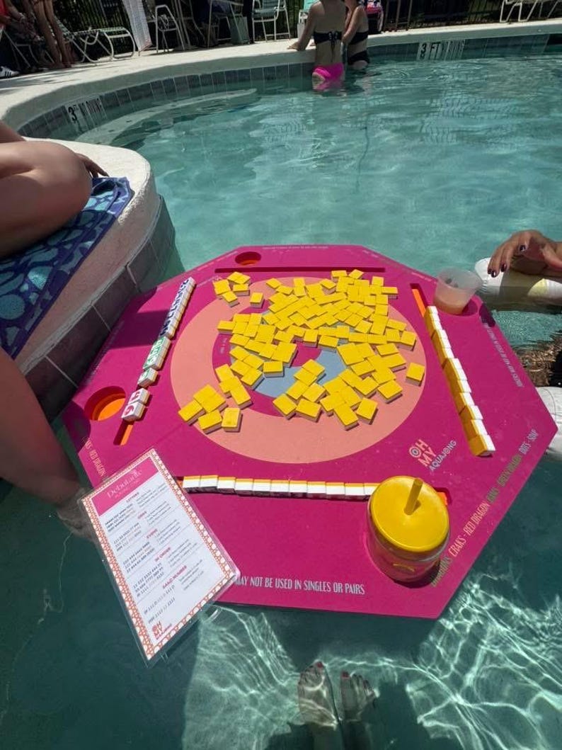 May include: A pink hexagonal floating game board with yellow tiles and game pieces. The board is in a swimming pool with people in the water. A menu is visible, along with the text "OH MY AQUAJONG".