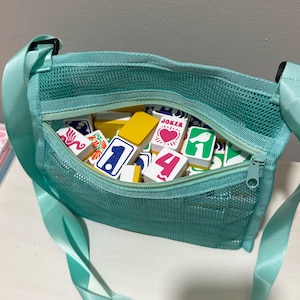 May include: A teal mesh crossbody bag filled with colorful game tiles. The bag has a zippered top and an adjustable strap. The tiles feature various numbers, symbols, and the word "JOKER".