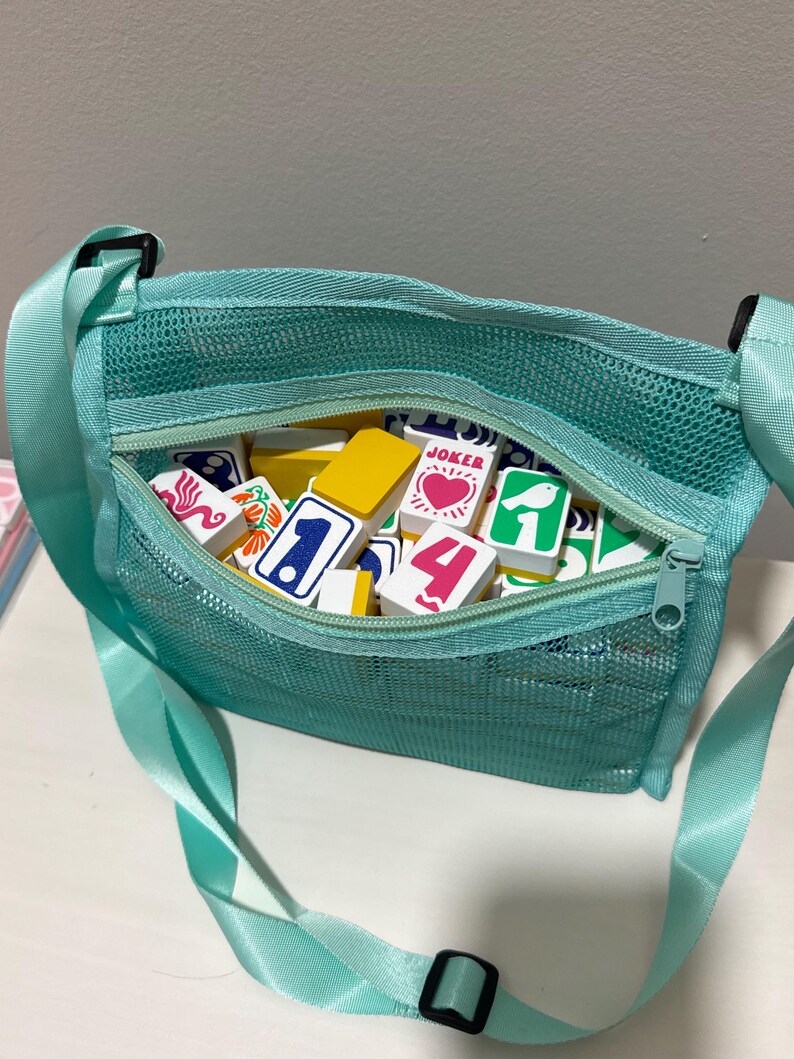 May include: A teal mesh crossbody bag filled with colorful game tiles. The bag has a zippered top and an adjustable strap. The tiles feature various numbers, symbols, and the word "JOKER".