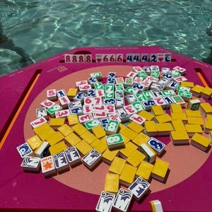 May include: A vibrant pink Mahjong game set with yellow tiles and various symbols and numbers. The tiles are scattered on a pink surface, with a background of a swimming pool. The game set includes tiles with the word "Joker".